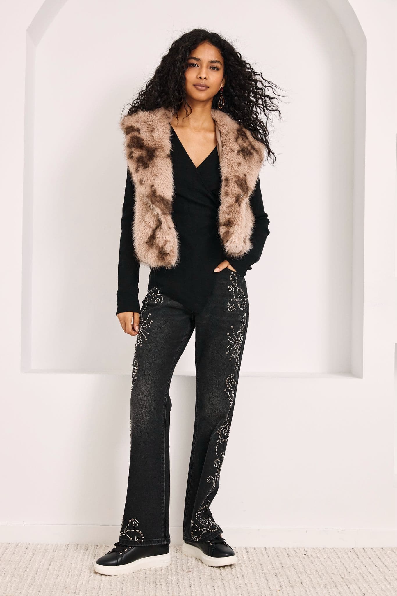 Model in a dark faux fur gilet over a black top and sequined trousers, studio background