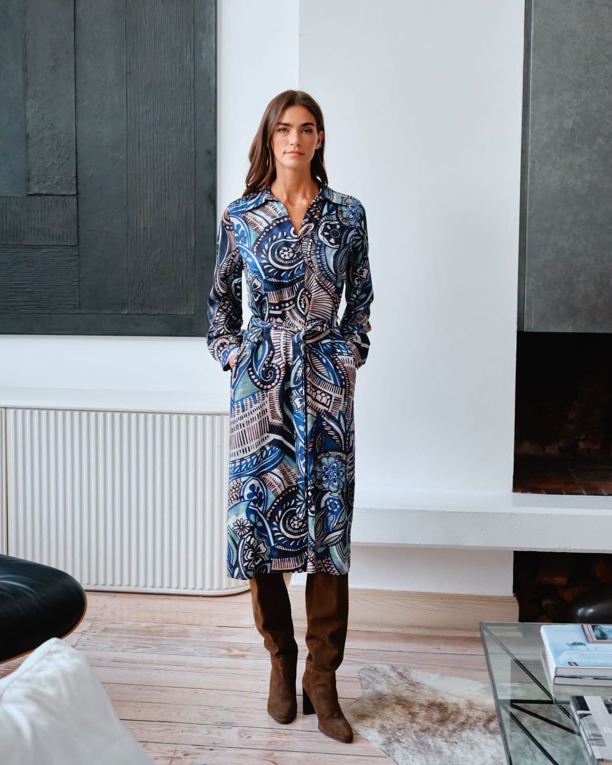 Model in a navy paisley-print wrap dress and boots, standing beside a window in a wood-floored interior