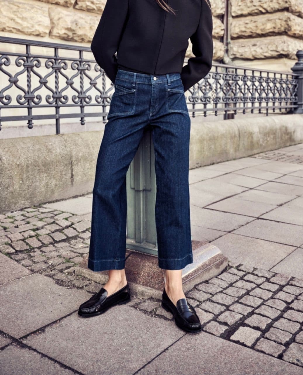 Model in a black top and wide-leg dark denim, standing by ornate street railings