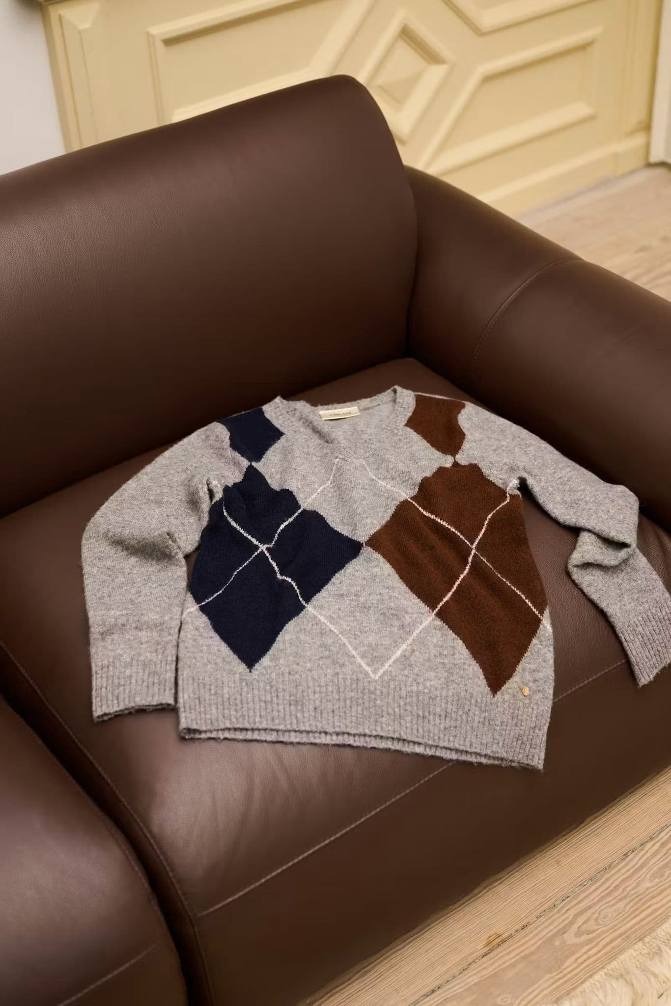 Grey argyle-pattern jumper with navy and brown diamonds, styled flat on a brown leather armchair