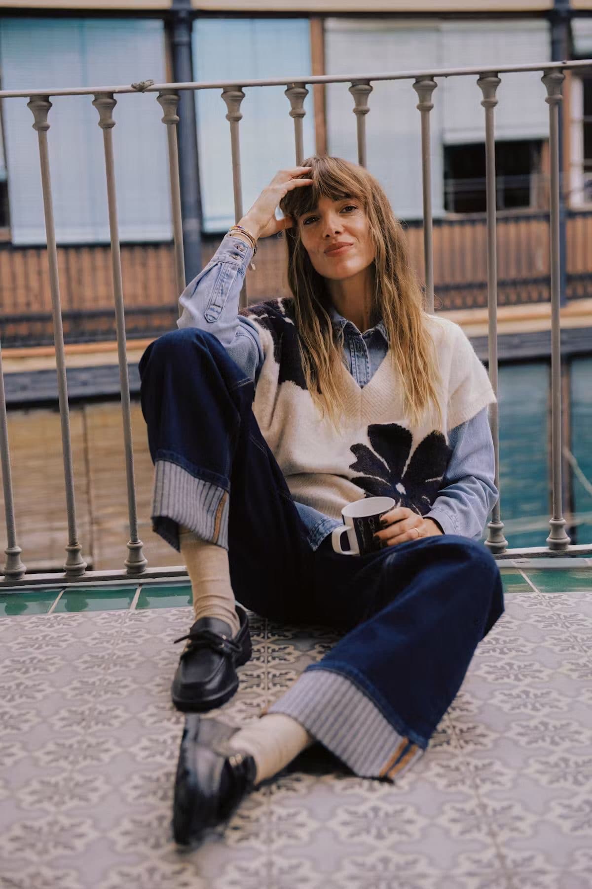 Model seated wearing a leaf-motif colourblock knit and wide-leg navy trousers on a street, wearing a cap